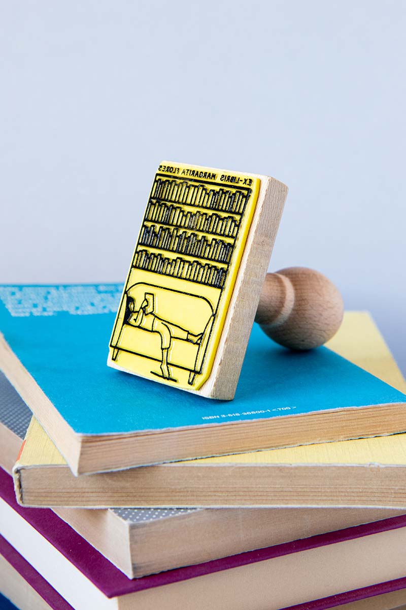 Sello ex-libris con una chica estirada leyendo en un sofa y una biblioteca detrás