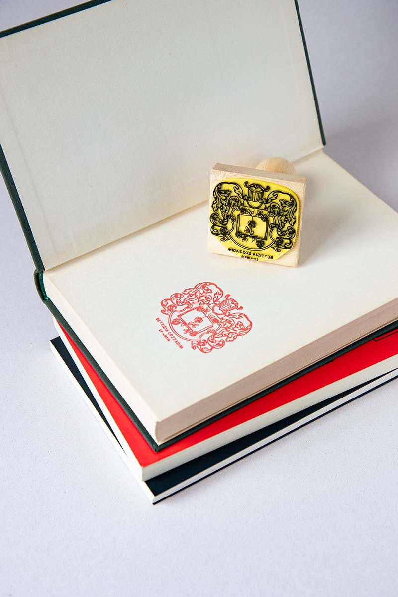 Sello ex-libris con con un escudo heráldico con yelmo en el interior hay un libro del que salen rosas