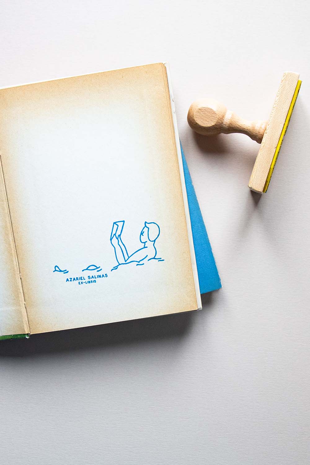 Portadilla de libro estampada con sello exlibris de una persona leyendo un libro mientras nada y sello
